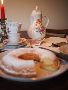 Französischer Apfelkuchen ohne Backpulver in Kranzform, unscharf im Hintergrund, im Vordergrund eine handgeschriebene Rezeptkarte neben einer floralen Kaffeekanne.