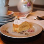 Französischer Apfelkuchen ohne Backpulver, als Stück auf einem floralen Porzellanteller serviert, mit Puderzucker bestäubt, daneben Besteck und Teeservice im Hintergrund.